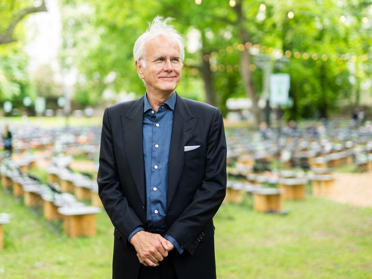 Harald Schmidt steht schmunzelnd und zur Seite schauend in einem Garten, im Hintergrund sind Sitzhocker zu sehen. Er trägt Brille, einen schwarzen Anzug und ein blaues Hemd.
