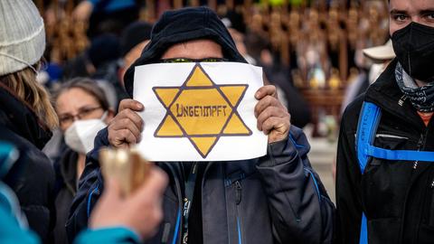 Demonstration gegen Coronamaßnahmen hält vor sein Gesicht ein Plakat auf dem ein gelber Davidstern abgebildet ist, in dessen Mitte steht "ungeimpft". 