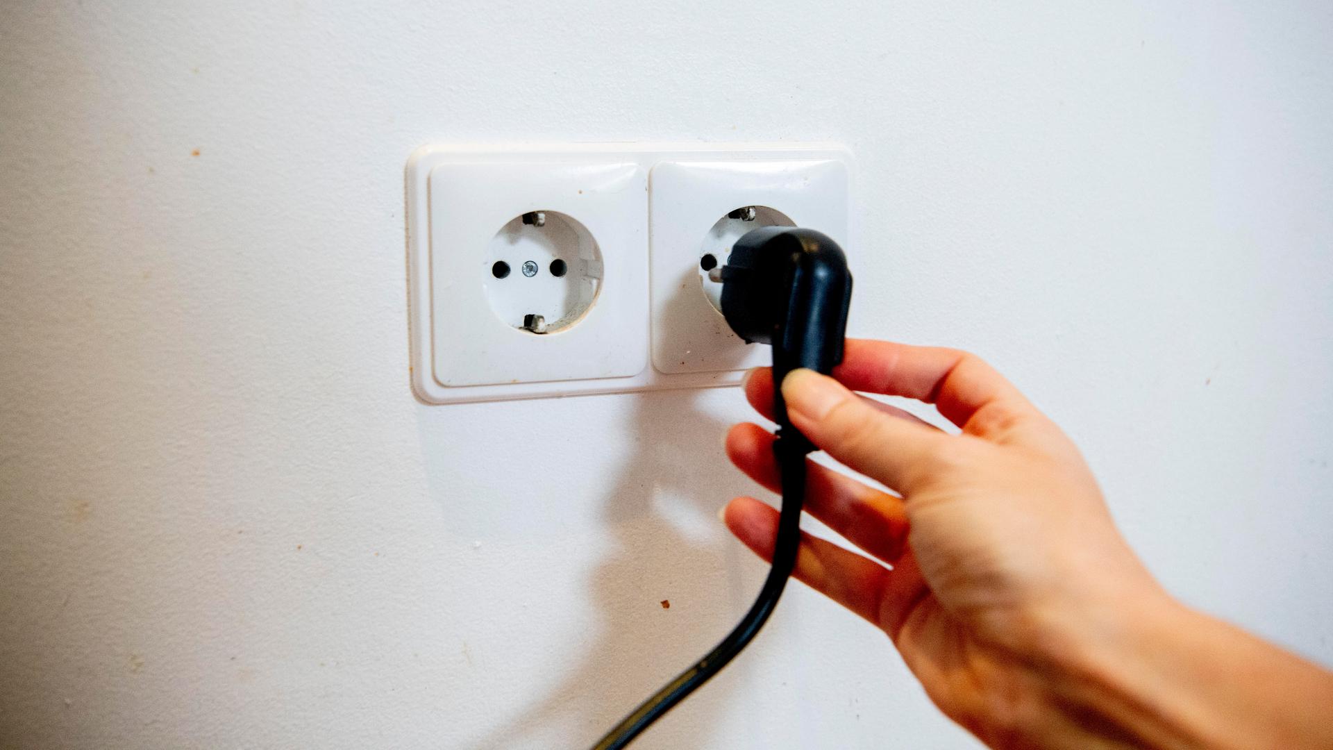 Das Bild zeigt eine Steck-Dose an einer Wand. Eine Hand steckt gerade einen Stecker in die Steck-Dose.