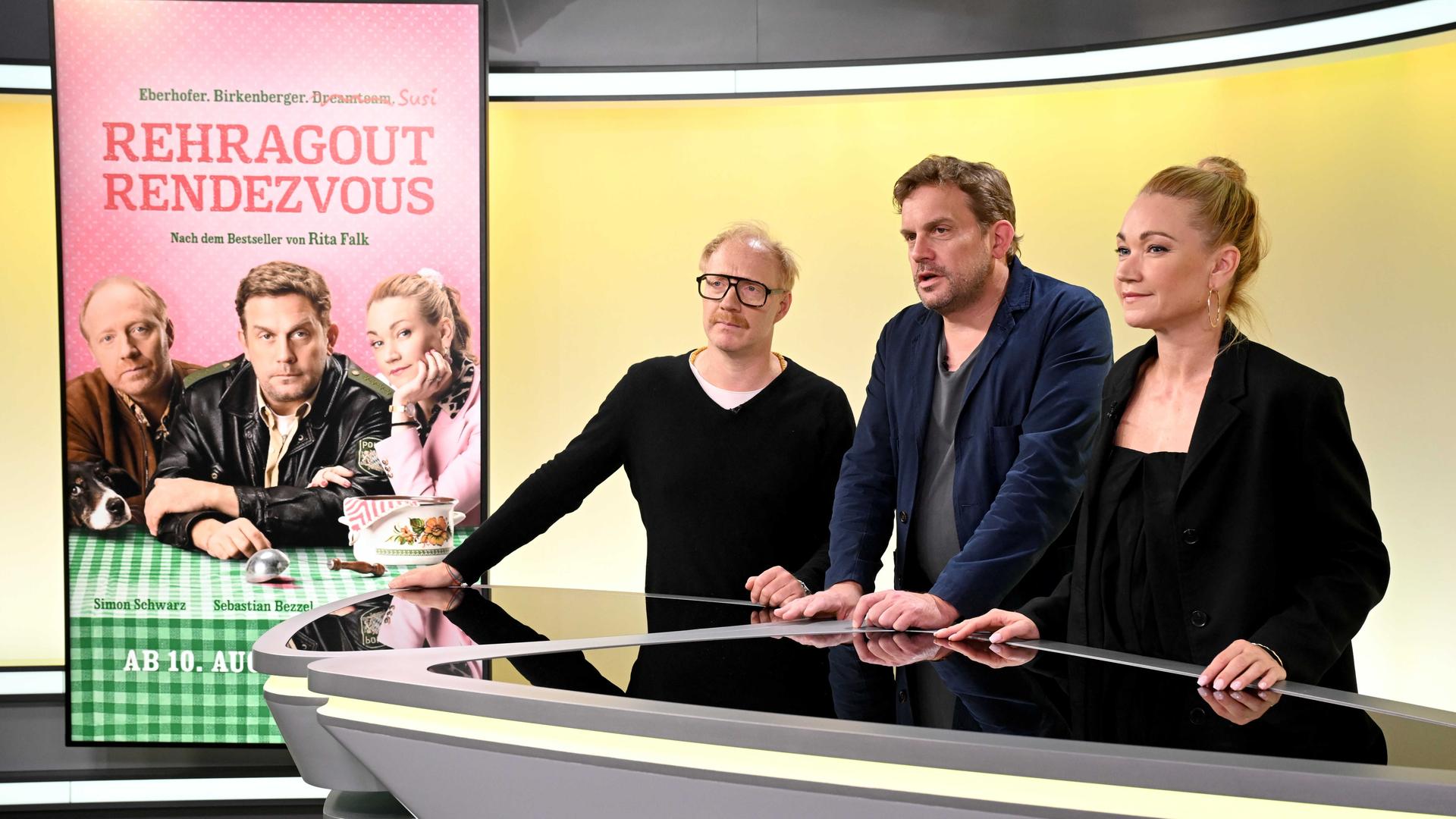 Von links nach rechts stehen die Schauspieler Simon Schwarz, Sebastian Bezzel und Lisa Maria Potthoff stehen in einem Studio an einem Tisch, links von ihnen ist ein Filmplakat zu sehen mit dem Titel "Rehragout-Rendezvous". 