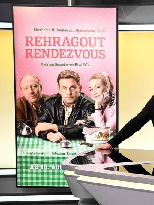 Von links nach rechts stehen die Schauspieler Simon Schwarz, Sebastian Bezzel und Lisa Maria Potthoff stehen in einem Studio an einem Tisch, links von ihnen ist ein Filmplakat zu sehen mit dem Titel "Rehragout-Rendezvous". 