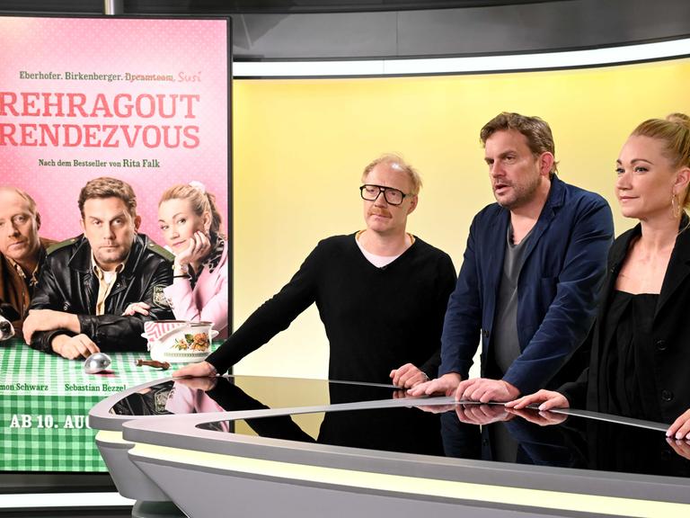 Von links nach rechts stehen die Schauspieler Simon Schwarz, Sebastian Bezzel und Lisa Maria Potthoff stehen in einem Studio an einem Tisch, links von ihnen ist ein Filmplakat zu sehen mit dem Titel "Rehragout-Rendezvous". 