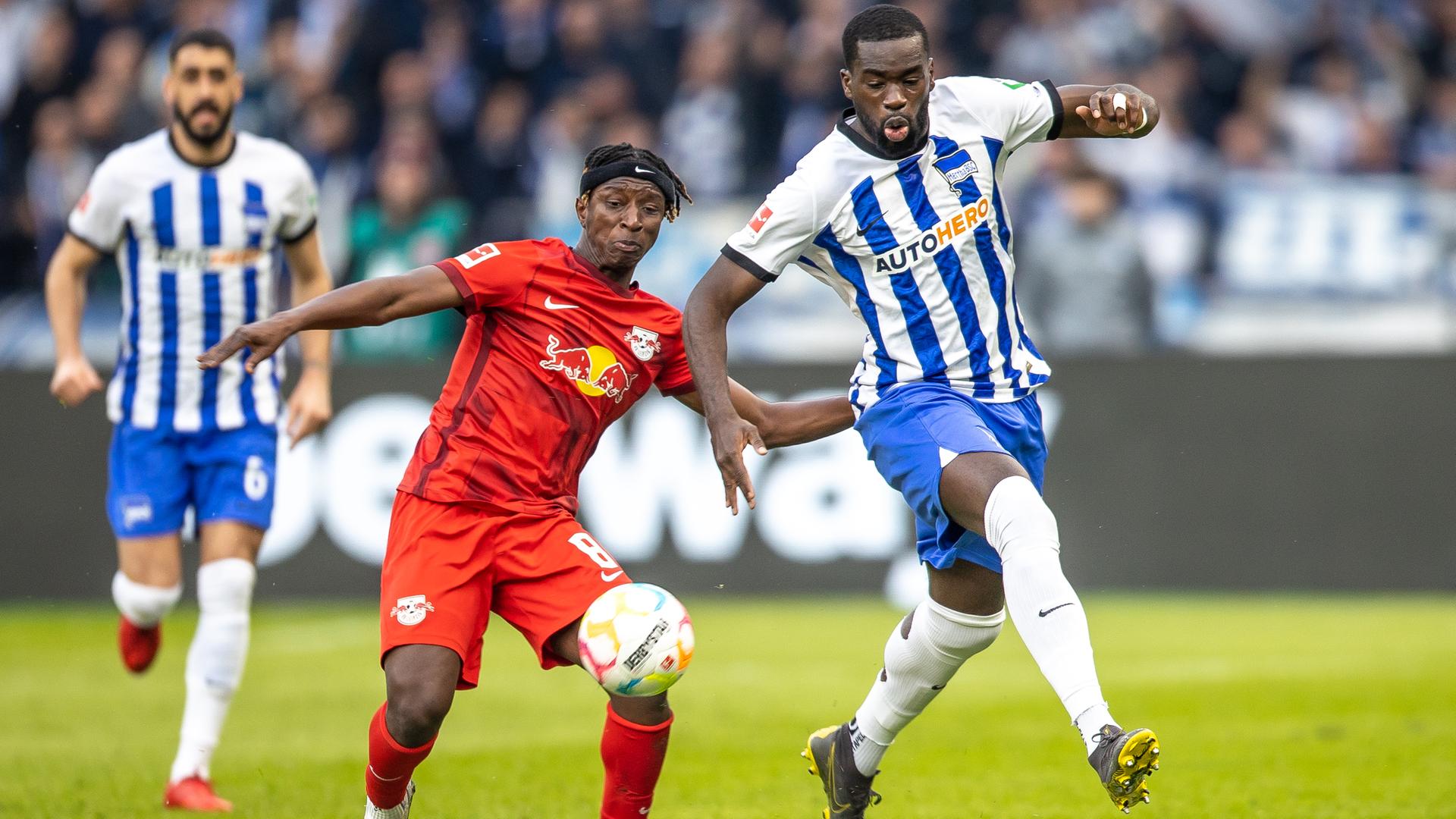 Die beiden rennen nebeneinander; vor ihnen fliegt der Ball. Haidara trägt ein rotes Trikot, Kanga ein blau-weiß-gestreiftes. Im Hintergrund unscharf ein weiterer Hertha-Spieler.