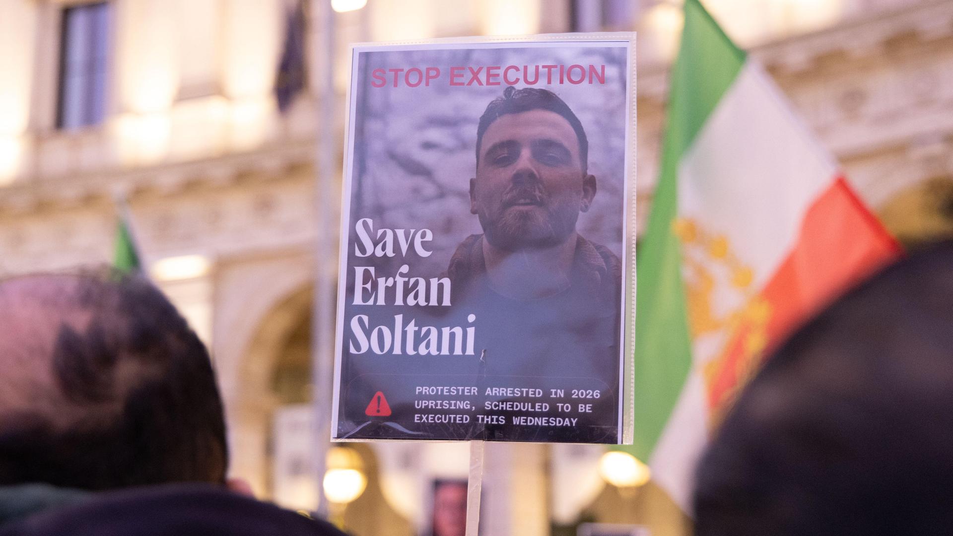 Das Bild wird mit einem Stock hochgehalten, auf dem der Oberkörper Soltanis zu sehen ist. Dahinter unscharf eine iranische Fahne und eine Gebäudefront. Im Vordergrund ebenfalls unscharf die Hinterköpfe von zwei Demonstranten.
