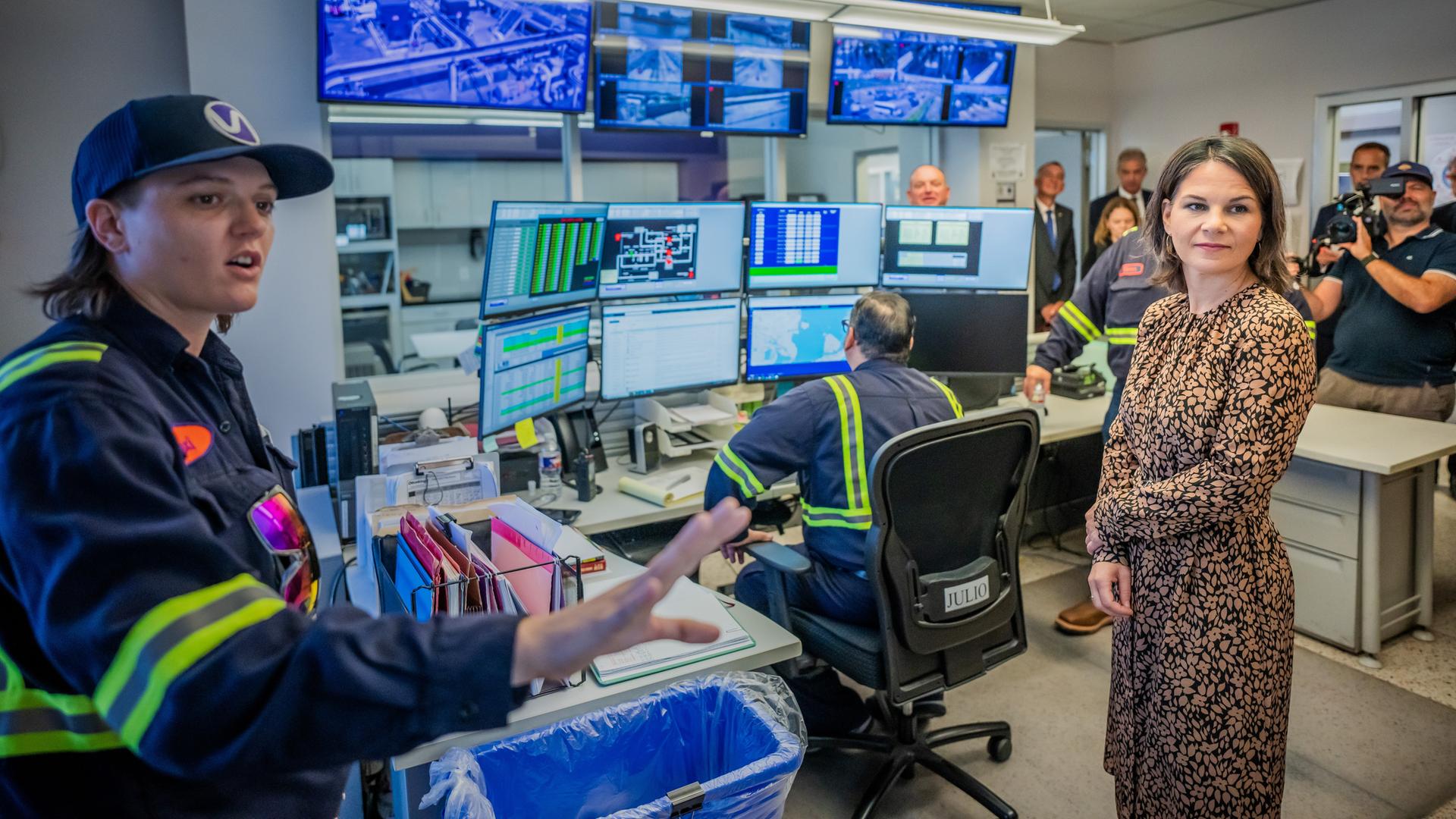 Baerbock im braun-schwarz gemusterten Kleid steht in einem Raum mit vielen Monitoren und Mitarbeitern. Eine Frau erklärt gerade etwas. Baerbock im braun-schwarz gemusterten Kleid steht in einem Raum mit vielen Monitoren und Mitarbeitern. Eine Frau erklärt gerade etwas.