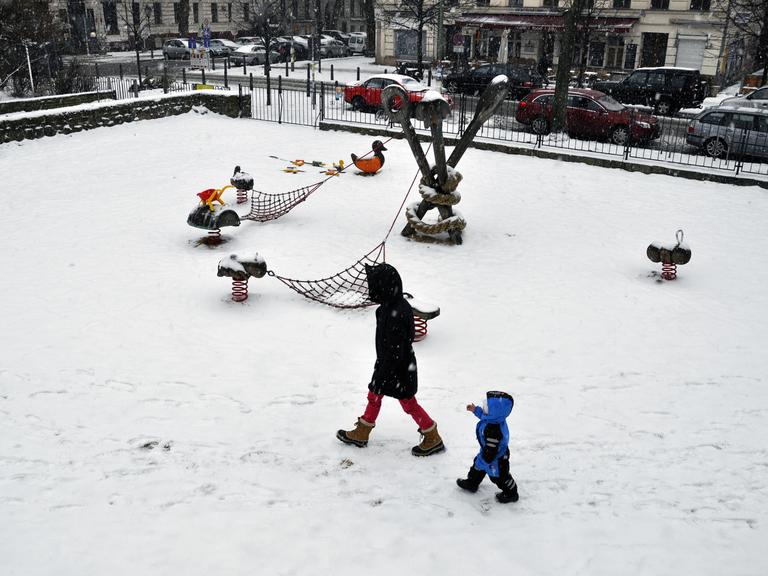 Eine Mutter mit ihrem Kind geht allein über einen verschneiter Spielplatz am Kollwitzplatz im Ortsteil Prenzlauer Berg in Berlin.