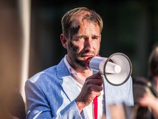 Der Aktionskünstler Philipp Ruch steht mit schwarz angemaltem Gesicht auf einer Straße und hält ein Megafon vor das Gesicht. Der Aktionskünstler Philipp Ruch steht mit schwarz angemaltem Gesicht auf einer Straße und hält ein Megafon vor das Gesicht.