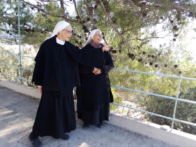Zwei ältere Damen im schwarzen Ornat katholischer Ordensschwestern befinden sich auf einem Gartenweg in angeregtem Gespräch, rechts von ihnen: ein Metallgeländer, von dem aus man ins Tal hinunterblicken kann.