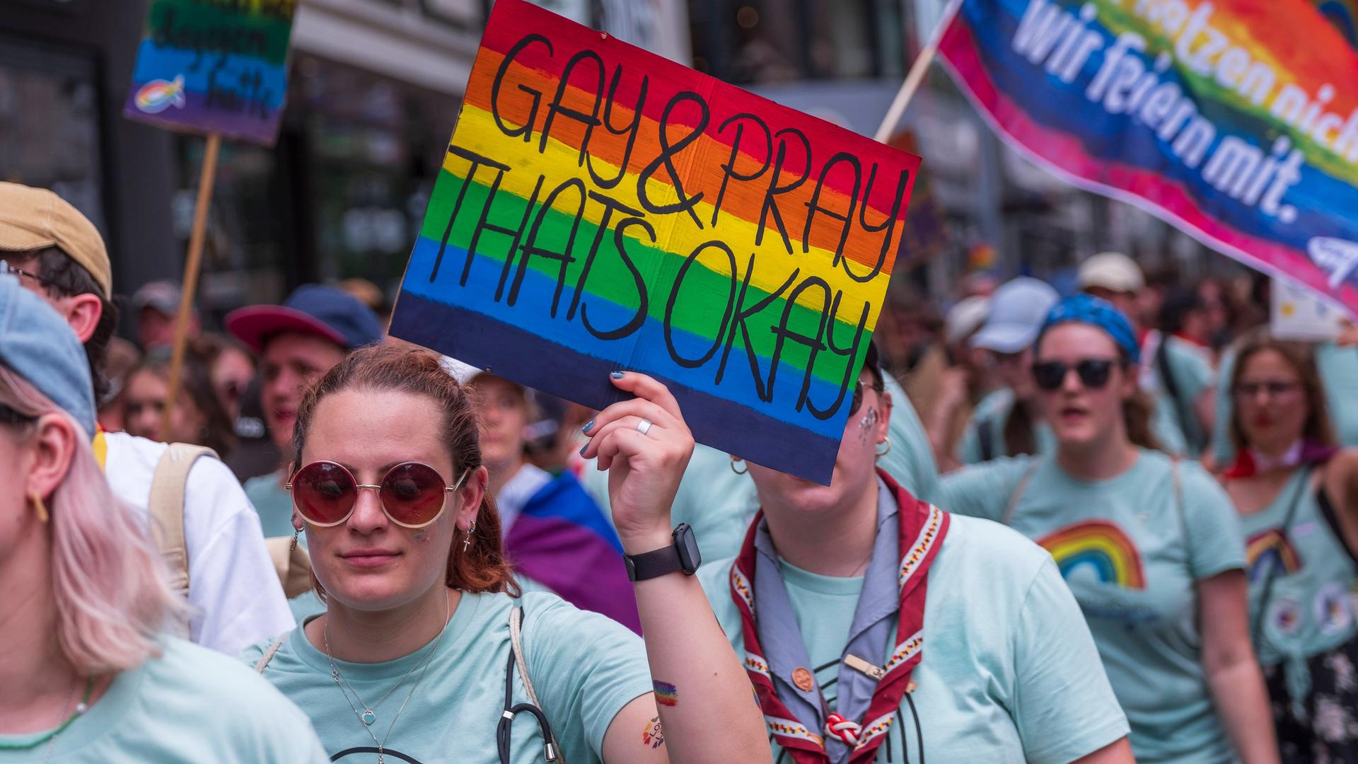 Eine weiblich gelesene Person mit Sonnenbrille hält im Vordergrund ein rechteckiges Schild in Regennbogenfarben in der linken Hand. Darauf steht: Gay & Pray Thats Okay. Sie ist Teil einer größeren Gruppe von Menschen, die die gleichen T-Shirts tragen. Sie laufen gemeinsam in Richtung der Kamera. Im Hintergrund trägt eine andere Person aus der Gruppe eine Regenbogenfahne, Darauf steht: Katholihsch. Wir hetzen nicht. Wir feiern mit. Ein weiteres Schild ist am linken Bildrand. Der Schriftzug ist allerdings unleserlich.