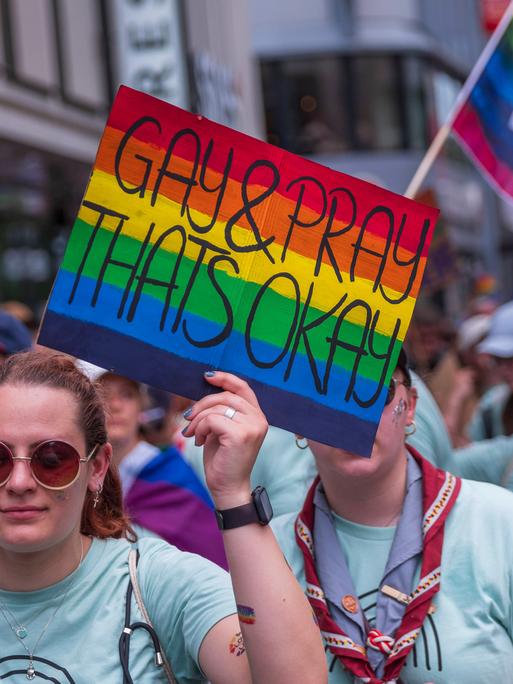 Eine weiblich gelesene Person mit Sonnenbrille hält im Vordergrund ein rechteckiges Schild in Regennbogenfarben in der linken Hand. Darauf steht: Gay & Pray Thats Okay. Sie ist Teil einer größeren Gruppe von Menschen, die die gleichen T-Shirts tragen. Sie laufen gemeinsam in Richtung der Kamera. Im Hintergrund trägt eine andere Person aus der Gruppe eine Regenbogenfahne, Darauf steht: Katholihsch. Wir hetzen nicht. Wir feiern mit. Ein weiteres Schild ist am linken Bildrand. Der Schriftzug ist allerdings unleserlich.