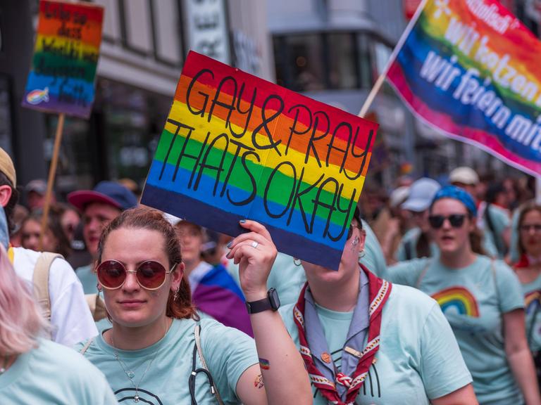 Eine weiblich gelesene Person mit Sonnenbrille hält im Vordergrund ein rechteckiges Schild in Regennbogenfarben in der linken Hand. Darauf steht: Gay & Pray Thats Okay. Sie ist Teil einer größeren Gruppe von Menschen, die die gleichen T-Shirts tragen. Sie laufen gemeinsam in Richtung der Kamera. Im Hintergrund trägt eine andere Person aus der Gruppe eine Regenbogenfahne, Darauf steht: Katholihsch. Wir hetzen nicht. Wir feiern mit. Ein weiteres Schild ist am linken Bildrand. Der Schriftzug ist allerdings unleserlich.