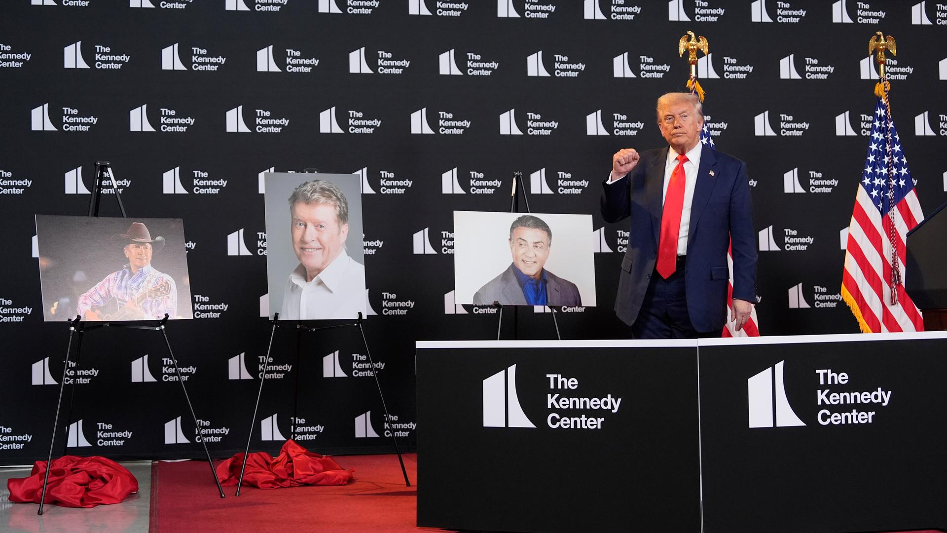 Trump steht vor einer Wand mit weißen Logos des Kennedy Centers. Links neben ihm stehen drei Bilder von Preisträgern auf Ständern. Hinter ihm zwei US-Fahnen. Trump gestikuliert mit der rechten Faust. 