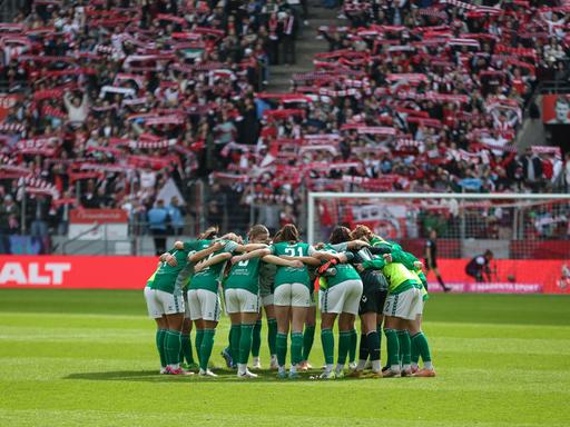 Die Spielerinnen des SV Werder Bremen schwören sich mit einem Mannschaftskreis im Stadion des 1. FC Kölns ein. Im Hintergrund halten Anhänger des 1. FC Köln Schals ihres Vereins in die Luft.