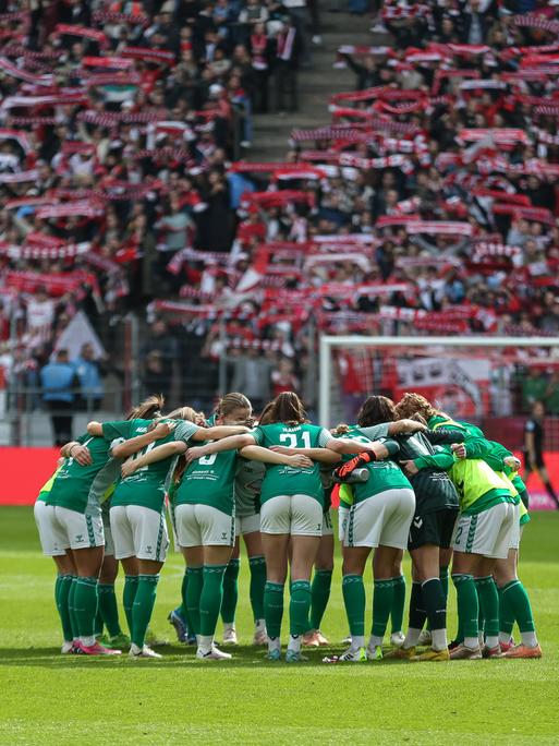 Die Spielerinnen des SV Werder Bremen schwören sich mit einem Mannschaftskreis im Stadion des 1. FC Kölns ein. Im Hintergrund halten Anhänger des 1. FC Köln Schals ihres Vereins in die Luft.