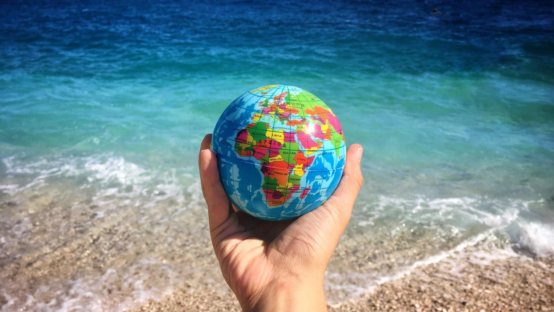Eine Hand hält einen kleinen Globus, im Hintergrund sieht man das Meer.