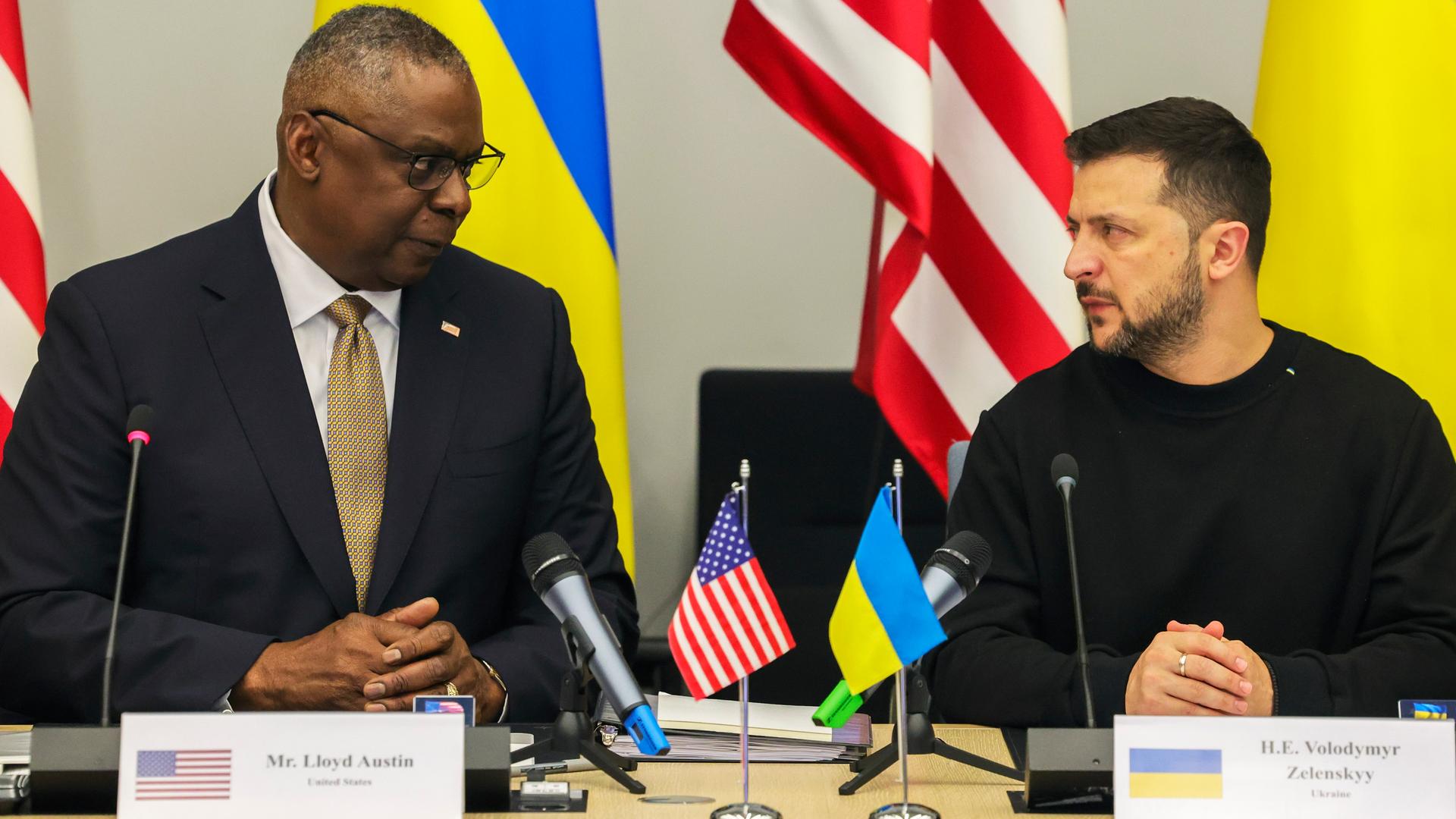 US-Verteidigungsminister Austin und der ukrainische Präsident Selenskyj sitzen nebeneinander vor Mikrofonen. Vor ihnen stehen ihre Namensschilder und die Landesfahnen.