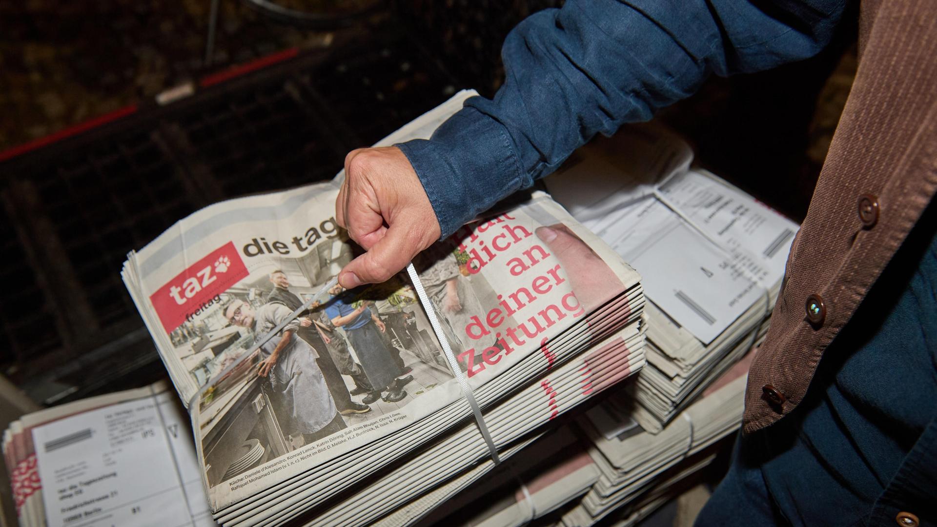 Eine Person greift einen abgepackten Stapel Zeitungen mit der letzten druckfrischen Ausgabe der werktäglichen "taz" vom 17.10.2025 