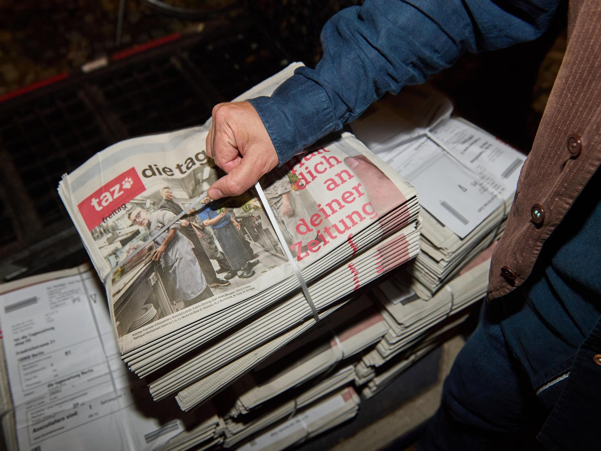 Eine Person greift einen abgepackten Stapel Zeitungen mit der letzten druckfrischen Ausgabe der werktäglichen "taz" vom 17.10.2025 