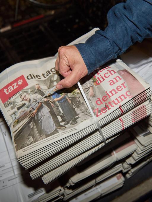 Eine Person greift einen abgepackten Stapel Zeitungen mit der letzten druckfrischen Ausgabe der werktäglichen "taz" vom 17.10.2025 