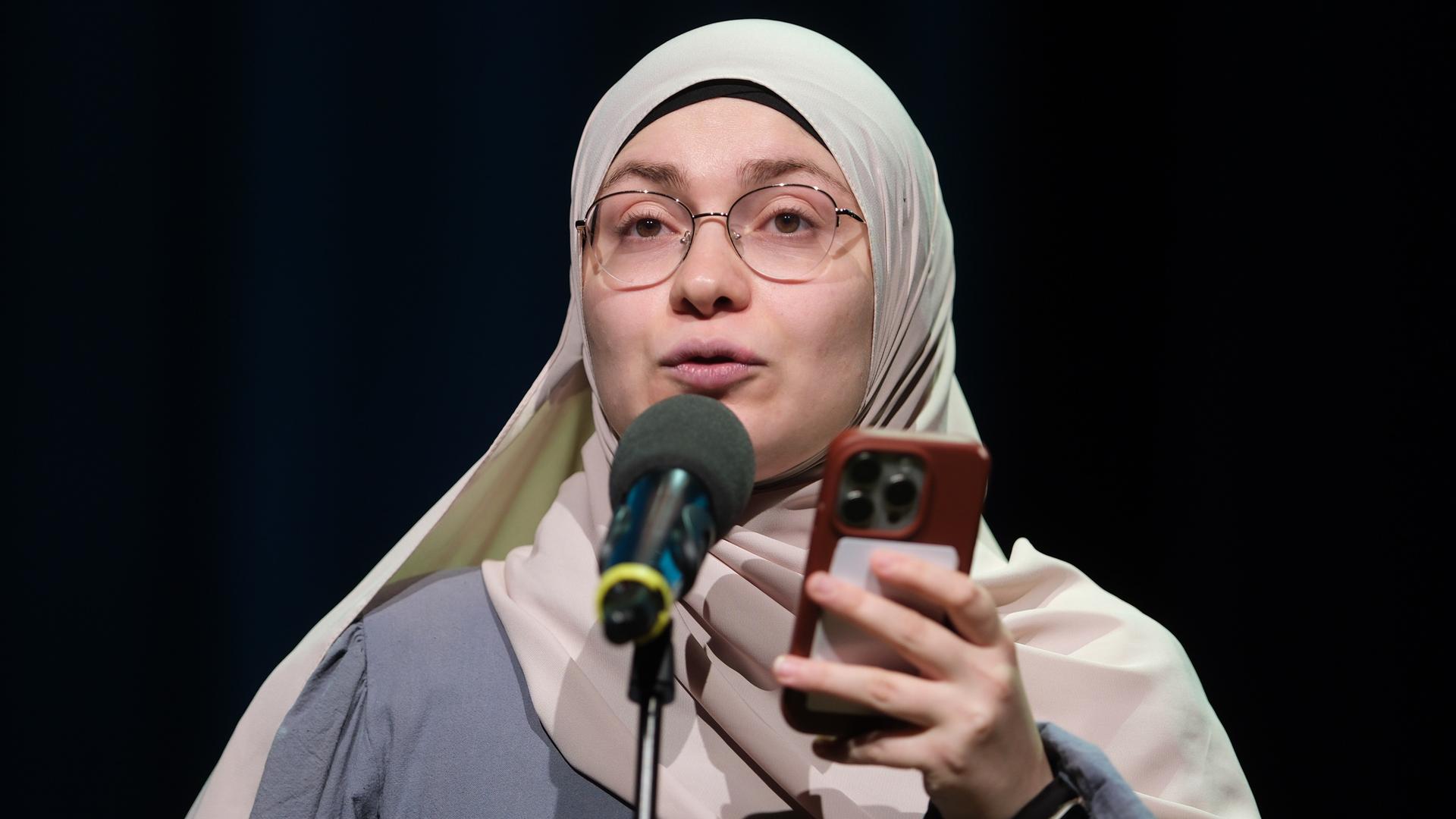 Ayse Irem steht hinter einem Mikrofon. Sie liest einen Text von ihrem Mobil-Telefon ab. 