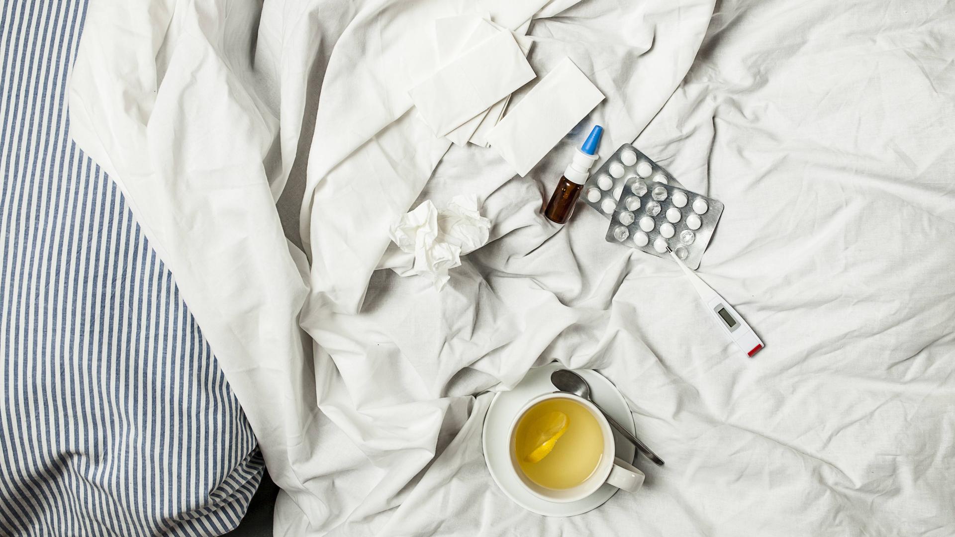 Zitronentee, Fieberthermometer, Tabletten, Nasenspray und benutzte Taschentücher liegen auf einem Bett.