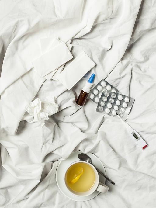 Zitronentee, Fieberthermometer, Tabletten, Nasenspray und benutzte Taschentücher liegen auf einem Bett.