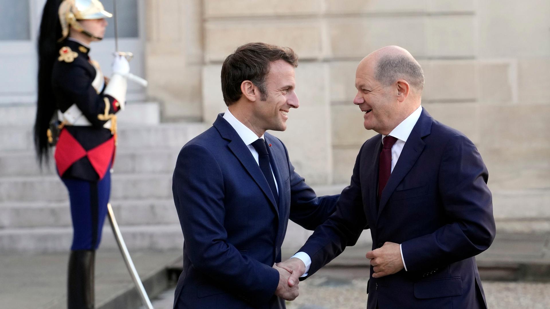 Der Präsident von Frankreich begrüßt den deutschen Bundes-Kanzler mit einem Hand-Schlag. Beide lachen sich an. Im Hinter-Grund steht eine Wache in Uniform.