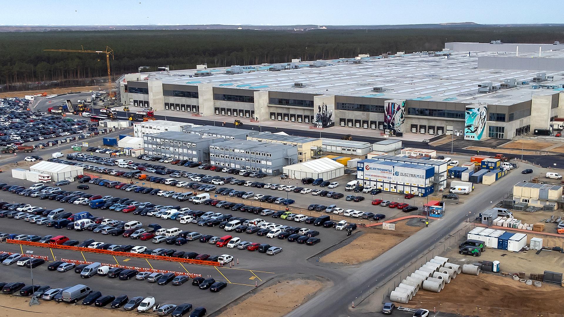 Außenansicht des Tesla-Werks in Grünheide: Vor einem langen Industriebau stehen hunderte Autos