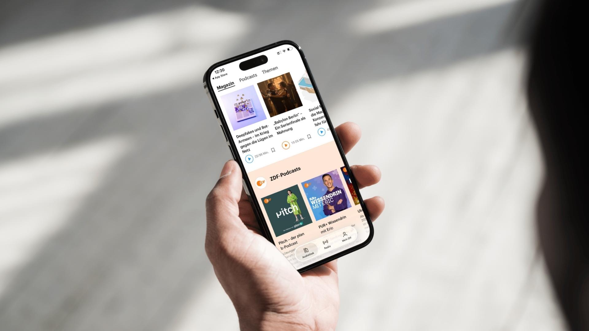 Eine Hand hält ein Smartphone auf dem die Oberfläche der Deutschlandfunk App mit integrierten ZDF-Podcasts zu sehen ist.