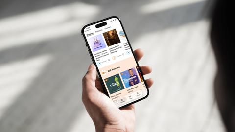 Eine Hand hält ein Smartphone auf dem die Oberfläche der Deutschlandfunk App mit integrierten ZDF-Podcasts zu sehen ist.