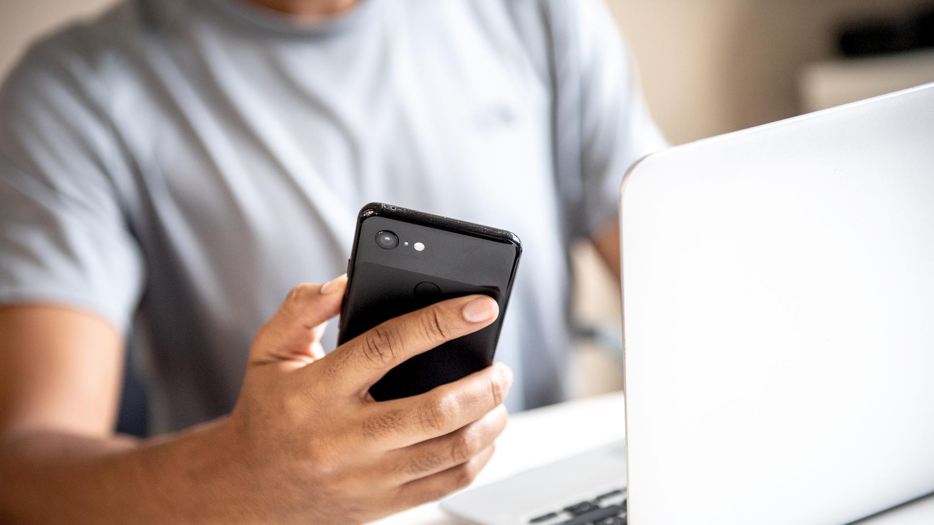 ILLUSTRATION - Ein junger Mann sitzt am 12.08.2019 in Berlin an einem Schreibtisch am Laptop und haelt ein Smartphone in der Hand (gestellte Szene). Foto: Zacharie Scheurer