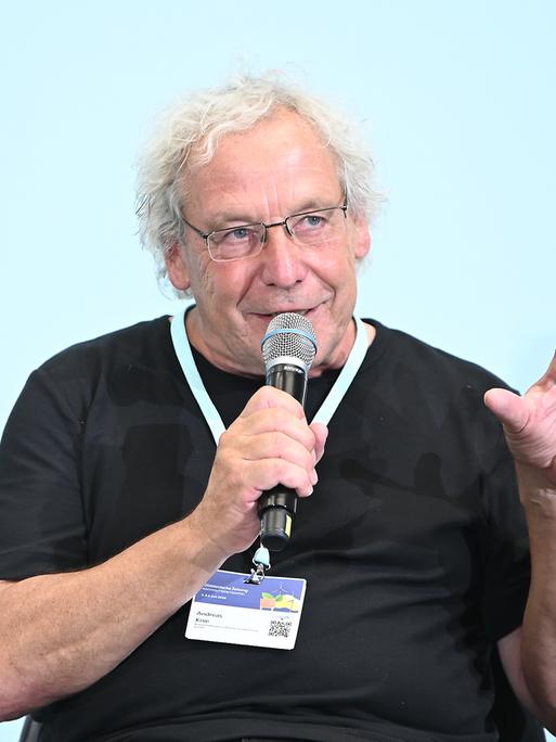 Der Soziologe und Verkehrsforscher Andreas Knie auf dem Nachhaltigkeitsgipfel 2025 bei einer Diskussion. Er trägt ein schwarzes T-Shirt, eine Brille und hat ein Mikrofon in der rechten Hand, in das er hinein spricht.