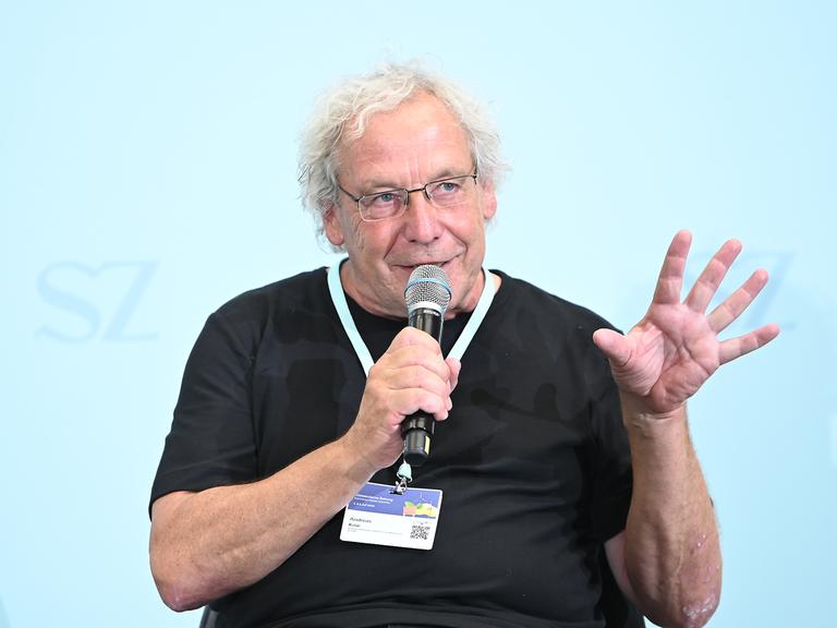 Der Soziologe und Verkehrsforscher Andreas Knie auf dem Nachhaltigkeitsgipfel 2025 bei einer Diskussion. Er trägt ein schwarzes T-Shirt, eine Brille und hat ein Mikrofon in der rechten Hand, in das er hinein spricht.