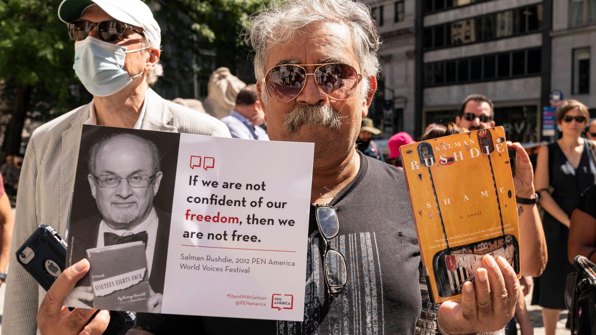 Ein Aktivist hält im August 2022 in New York während der PEN America-Kundgebung zur Unterstützung von Salman Rushdie ein Plakat mit einem Bild von Salman Rushdie und dem Zitat "If we are not confident of our freedom, then we are not free." sowie ein Buch von Rushdie.