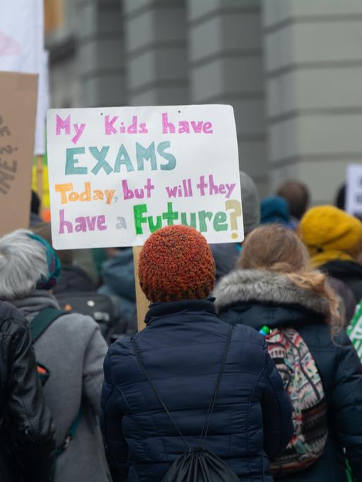 Eine Frau hält ein selbstgemaltes Plakat hoch. Darauf steht auf Englisch: „My kids have EXAMS today, but will they have a FUTURE?“ Im Hintergrund sind weitere Demonstrierende zu sehen, einige mit bunten Schildern.