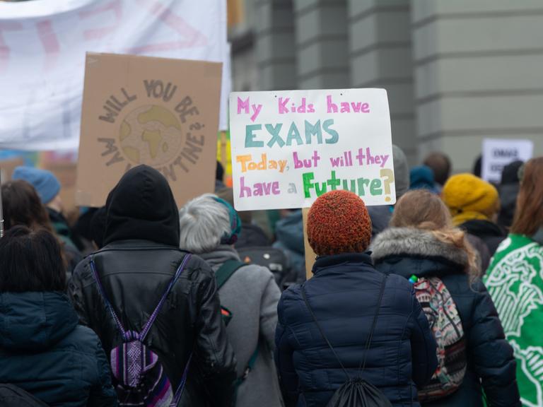 Eine Frau hält ein selbstgemaltes Plakat hoch. Darauf steht auf Englisch: „My kids have EXAMS today, but will they have a FUTURE?“ Im Hintergrund sind weitere Demonstrierende zu sehen, einige mit bunten Schildern.