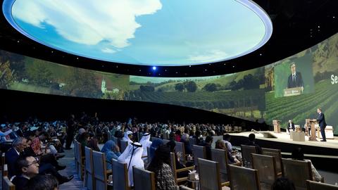 In einer Halle sitzen mehrere Vertreter im Publikum während auf der Bühne gesprochen wird. Die Decke der Halle ist mit einem riesigen Kreis perforiert, sodass der Himmel zu sehen ist.