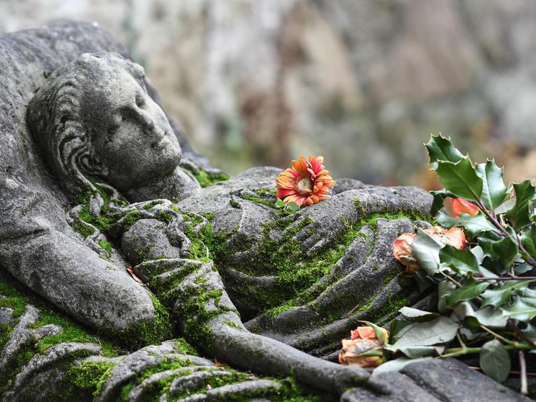 Mit Moos bewachsenes Steinrelief auf einem Grabmal zeigt eine schlafende junge Frau. Auf der Brust der Figur und neben ihr liegen frische Blumen.