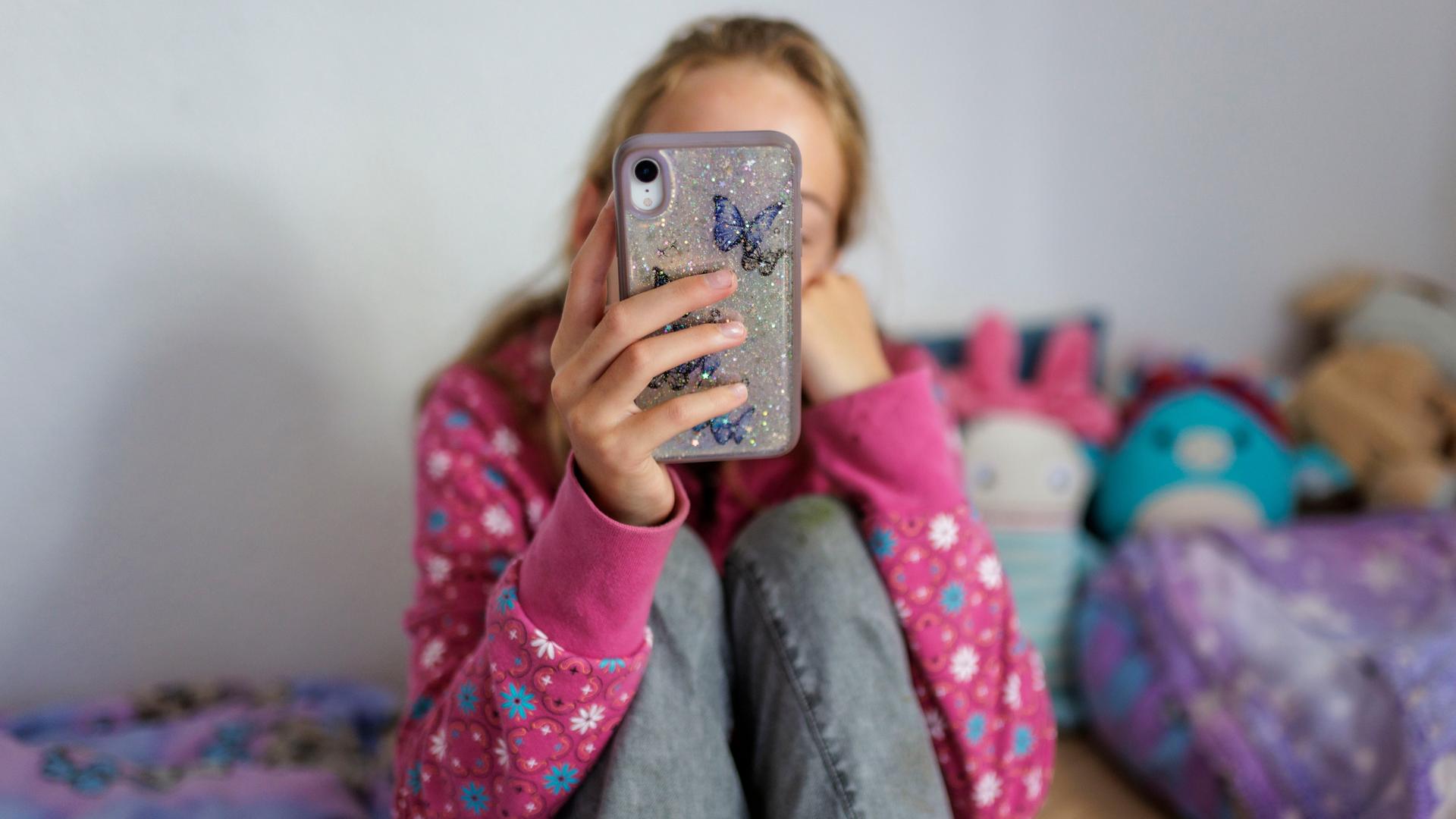 Ein Mädchen sitzt auf ihrem Bett und hält ein Handy vor ihr Gesicht (Symbolfoto)