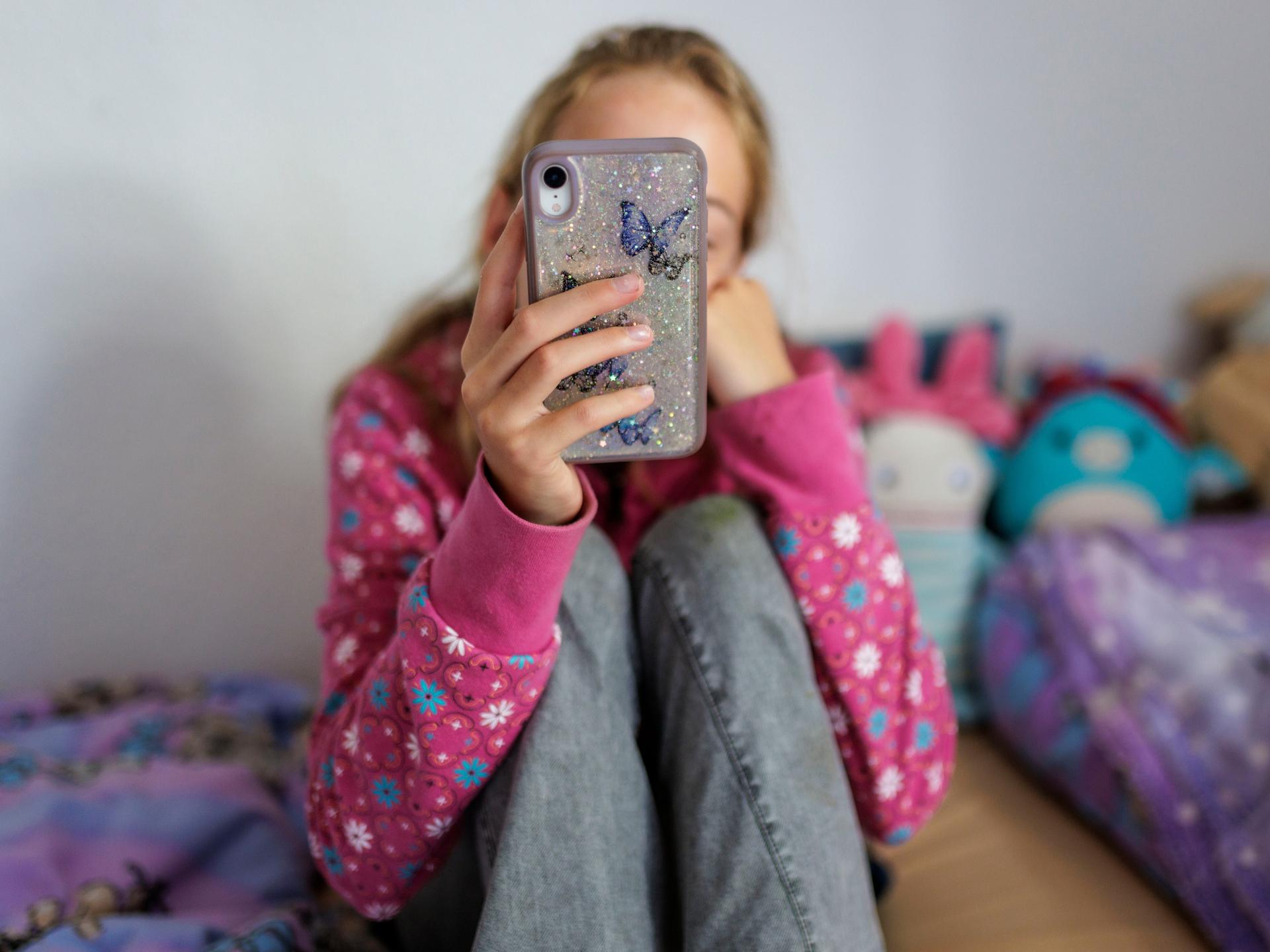 Ein Mädchen sitzt auf ihrem Bett und hält ein Handy vor ihr Gesicht (Symbolfoto)