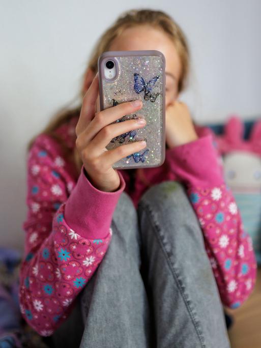 Ein Mädchen sitzt auf ihrem Bett und hält ein Handy vor ihr Gesicht (Symbolfoto) Ein Mädchen sitzt auf ihrem Bett und hält ein Handy vor ihr Gesicht (Symbolfoto)