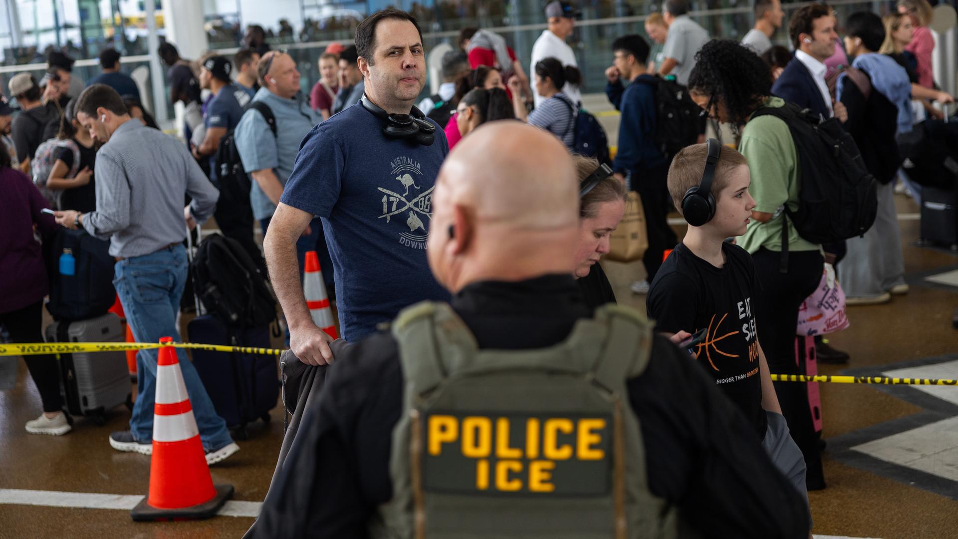 Ein uniformierter ICE-Agent steht an einem Flughafen und beobachtet die Menschen in einer Einreiseschlange. Ein uniformierter ICE-Agent steht an einem Flughafen und beobachtet die Menschen in einer Einreiseschlange.