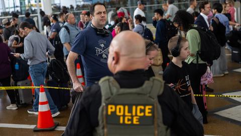 Ein uniformierter ICE-Agent steht an einem Flughafen und beobachtet die Menschen in einer Einreiseschlange.