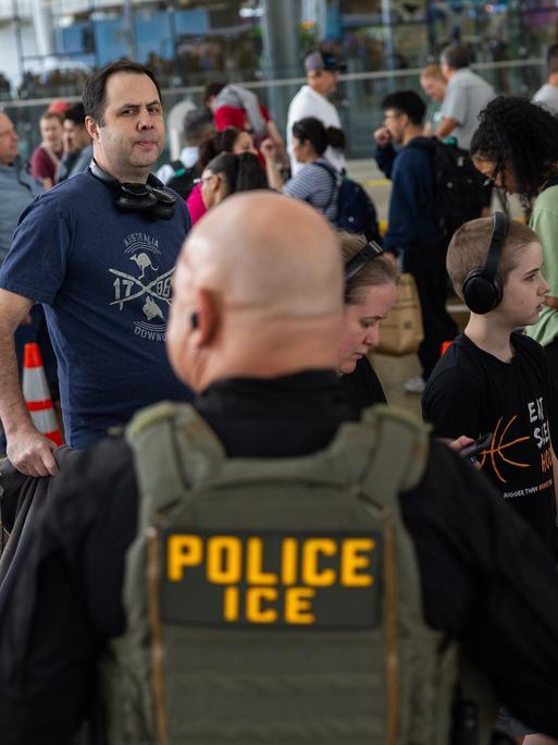 Ein uniformierter ICE-Agent steht an einem Flughafen und beobachtet die Menschen in einer Einreiseschlange.