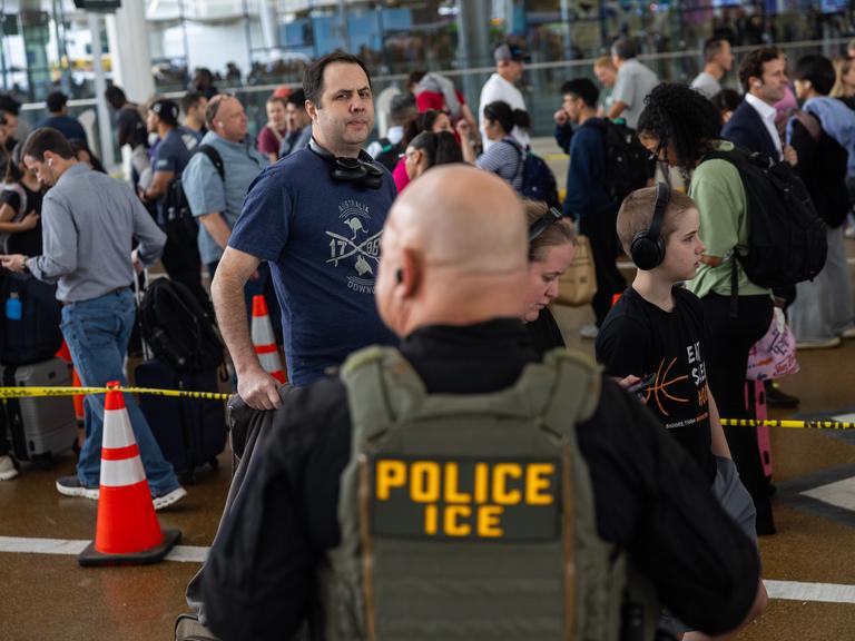 Ein uniformierter ICE-Agent steht an einem Flughafen und beobachtet die Menschen in einer Einreiseschlange.