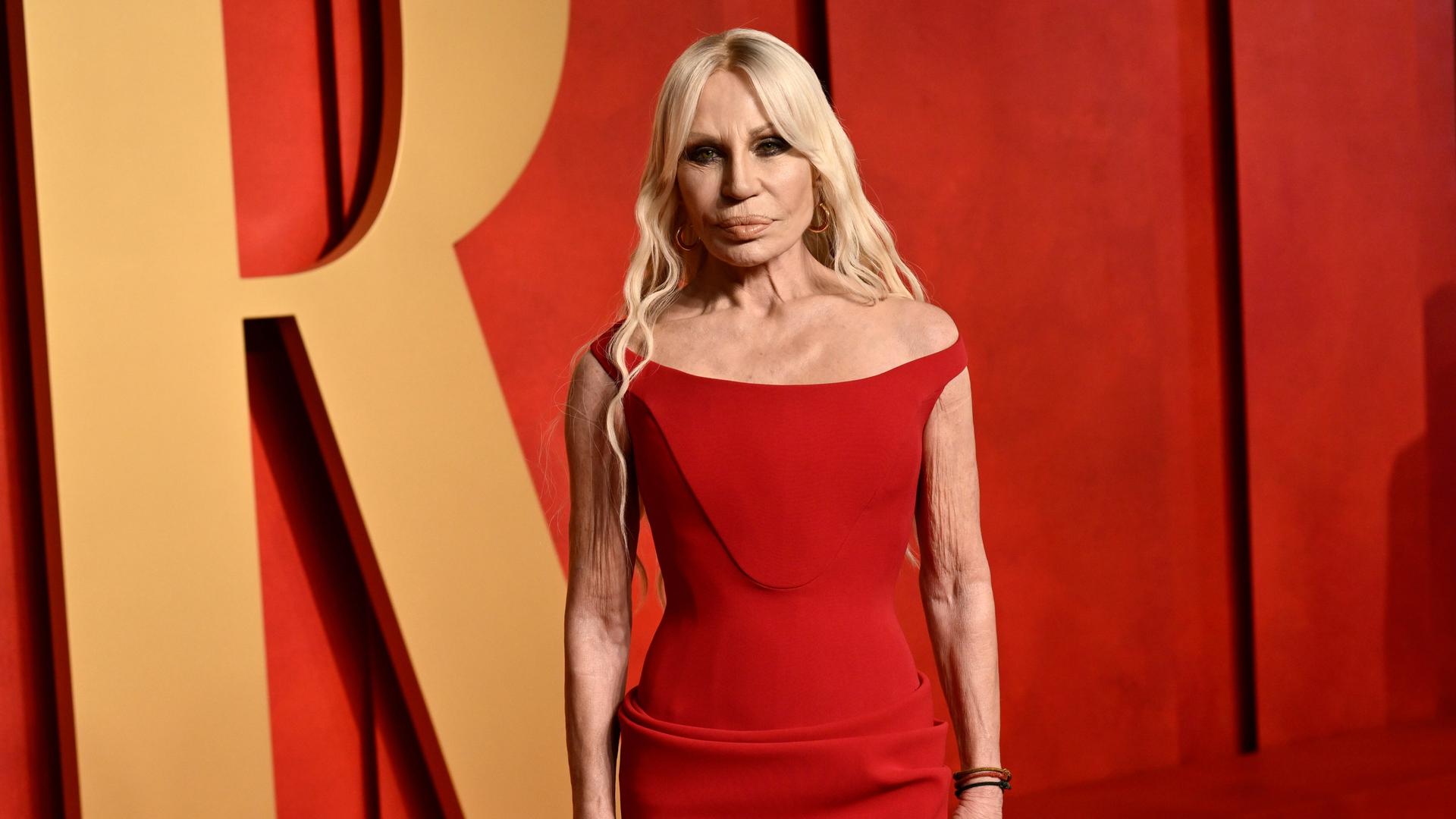 Die blondierte Donatella Versace im ärmellosen roten Kleid vor einer roten Wand mit einem großen bronzefarbenen "R".