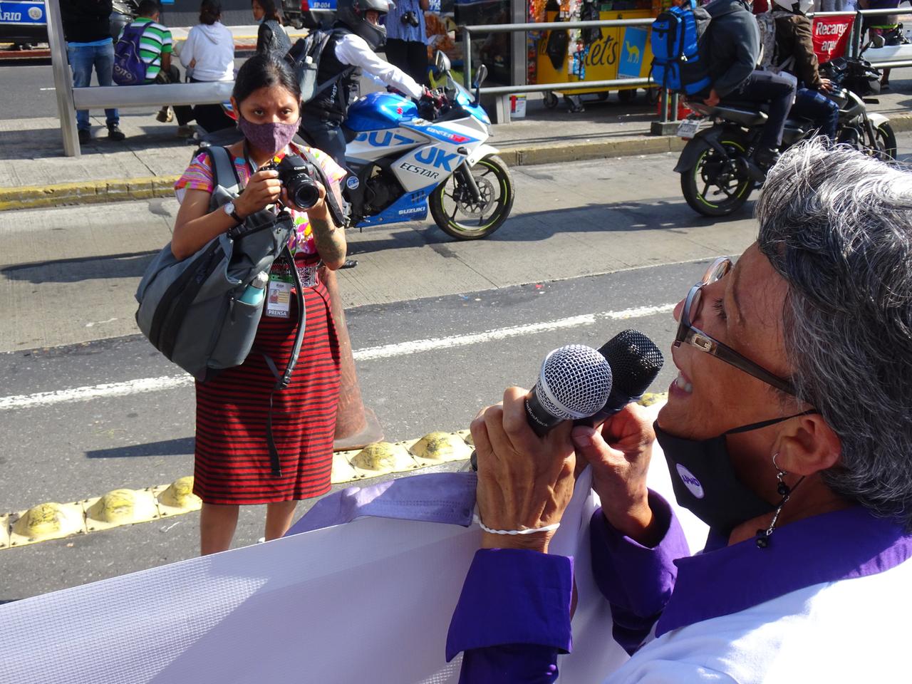 Eine Frau steht auf der Straße und richtet ihren Fotoapparat auf eine Frau im Vordergrund, die ein TRansparent hält und in zwei Mikrofone spricht.