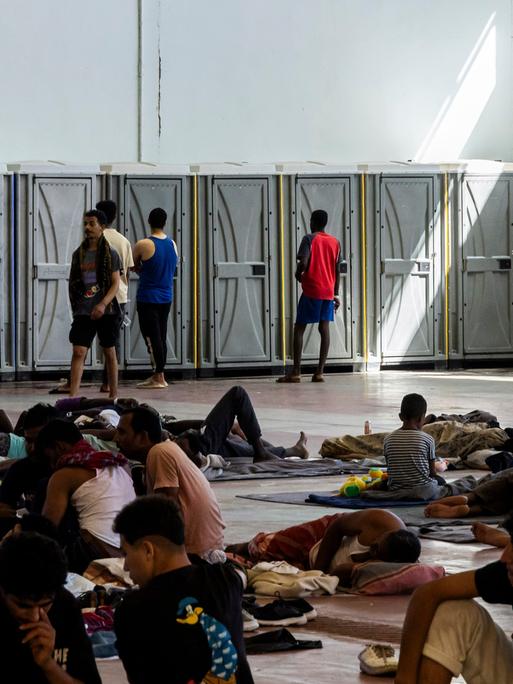 In einer Halle sind Dixie-Toiletten an einer Wand aufgestellt. Davor stehen Menschen. Außerdem liegen davor Menschen, teilweise nur auf Kartons. Es ist eine Flüchtlingsunterkunft auf der Insel Kreta.