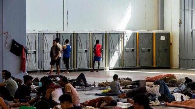 In einer Halle sind Dixie-Toiletten an einer Wand aufgestellt. Davor stehen Menschen. Außerdem liegen davor Menschen, teilweise nur auf Kartons. Es ist eine Flüchtlingsunterkunft auf der Insel Kreta.