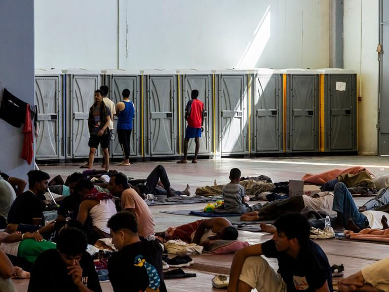 In einer Halle sind Dixie-Toiletten an einer Wand aufgestellt. Davor stehen Menschen. Außerdem liegen davor Menschen, teilweise nur auf Kartons. Es ist eine Flüchtlingsunterkunft auf der Insel Kreta.